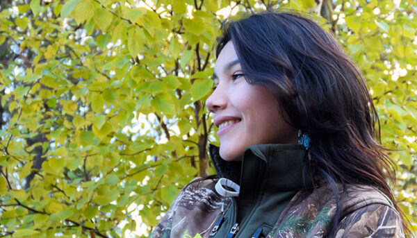 Jeune personne souriant en devant d'un arbre avec des feuilles verts et jaunes